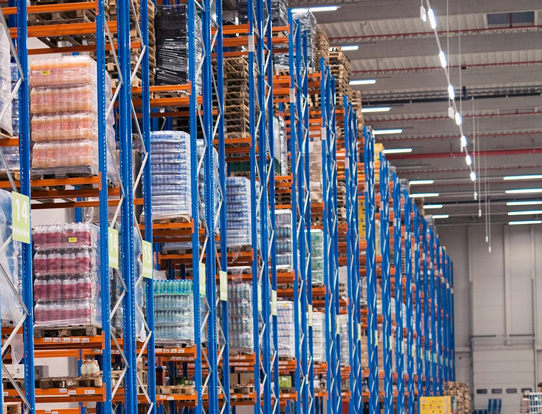 Interior of a large, well-organized warehouse with tall blue and orange shelving racks stocked with palletized goods—highlighting Facility Experts' capabilities in streamlining operations for industrial and logistics facilities.