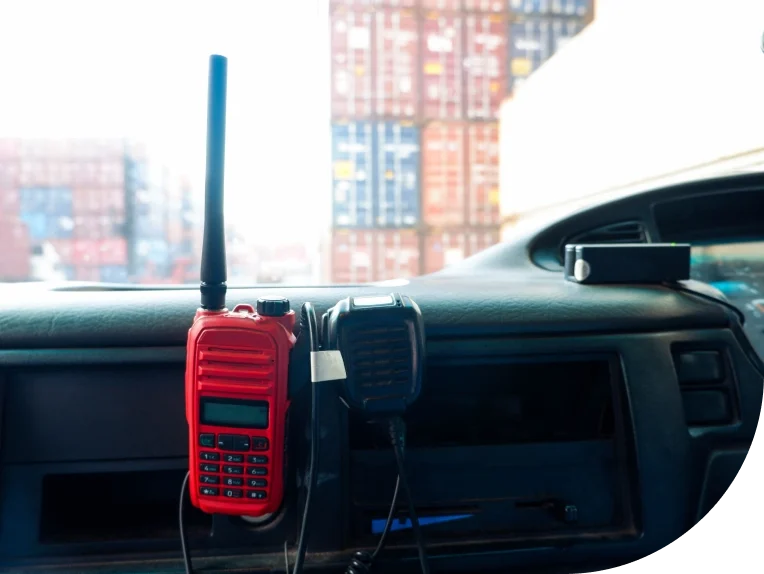 Red two-way radio mounted on a security vehicle dashboard with shipping containers visible through the windshield, illustrating mobile patrol operations.