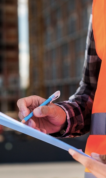 Worker in a safety vest writing on a clipboard at a job site, symbolizing compliance documentation.